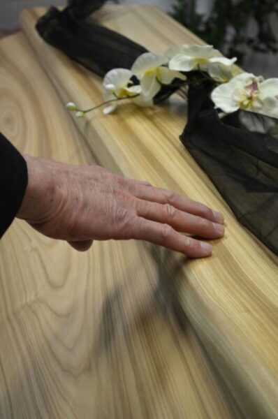 Close-up of a hand touching a wooden coffin adorned with flowers and black fabric, conveying a sense of mourning.